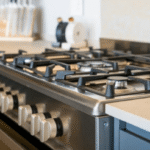 Stainless steel gas range with six burners and cream-colored control knobs beneath a light kitchen countertop.