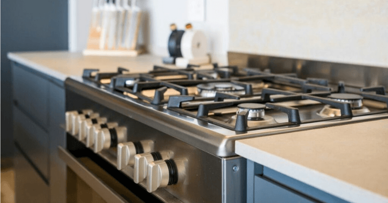 Stainless steel gas range with six burners and cream-colored control knobs beneath a light kitchen countertop.