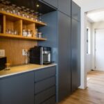 A modern kitchen featuring grey cabinetry, wooden shelves with glassware, and a coffee maker, with a light wood floor and a doorway leading to another room.