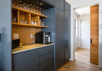 A modern kitchen featuring grey cabinetry, wooden shelves with glassware, and a coffee maker, with a light wood floor and a doorway leading to another room.